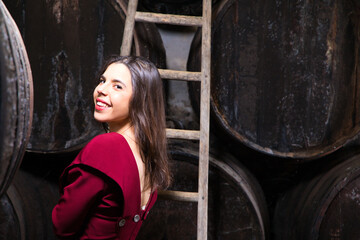 Fototapeta na wymiar Young, beautiful businesswoman posing in her wine cellar in front of a wooden wine barrel. The woman is wearing a red dress. Concept young and enterprising woman.