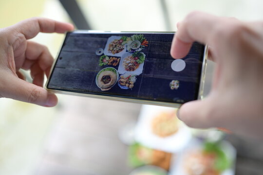 Woman holding a smartphone takes pictures of set Thai dishes and shares it on social networks before eating. Food served on wooden table consists of Wrapped in coconut, papaya salad,fried chicken,etc.