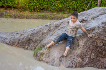Seven-year-old Asian boy plays mud slides having fun in muddy dirty clothes playing in school summer camp.