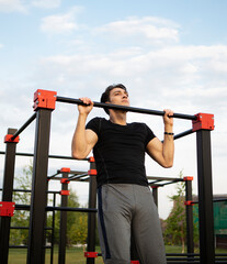 Front view of sporty man doing pull ups on horizontal bar outdoors. Young man street workout exercising at the morning time