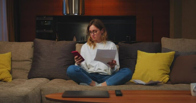 Female homeowner tenant puts on glasses to pay electricity bills, using mobile phone app siting on sofa at home 