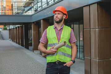   European engineer handsome man or architect holds a plan and a helmet, looking forward with orange safety helmet in construction site. Standing at modern building construction. 