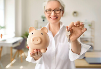 Happy smiling middle aged woman holding a pink piggy bank and house keys. Soft focus, close up....