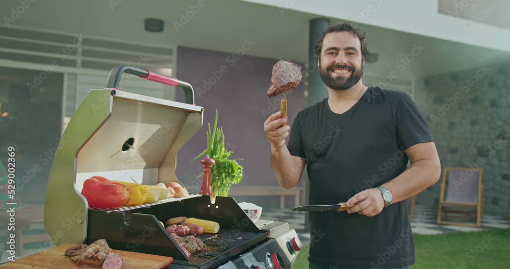 Wall mural Young man preparing meat on grill during summer garden party barbecue. BBQ chef showing steak food to camera