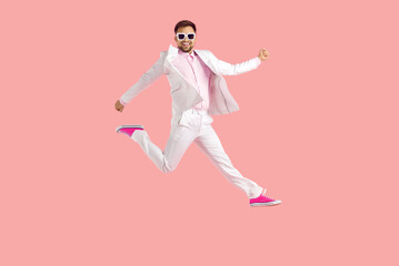 Funny confident young man having fun in the studio. Full body shot of happy excited joyful guy wearing white suit, eyeglasses and trainer shoes jumping high in the air isolated on pink background - Powered by Adobe