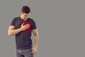 Fototapeta na wymiar Man suffering from heart disease. Young Caucasian male standing isolated on gray text copyspace background feels sudden pain and touches his chest in area marked with red color. Cardiac pain concept