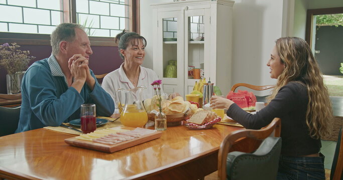 Happy Couple Having Breakfast With Teenage Daughter Sitting At Kitchen Table. The Father Is Talking And The Mother Is Serving Juice To The Daughter