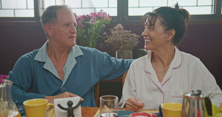 Middle aged couple having breakfast together. Older couple wearing pajamas talking and smiling. Senior woman drinking coffee. husband drinking orange juice.