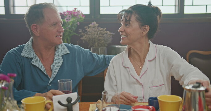 Middle Aged Couple Having Breakfast Together. Older Couple Wearing Pajamas Talking And Smiling. Senior Woman Drinking Coffee. Husband Drinking Orange Juice.