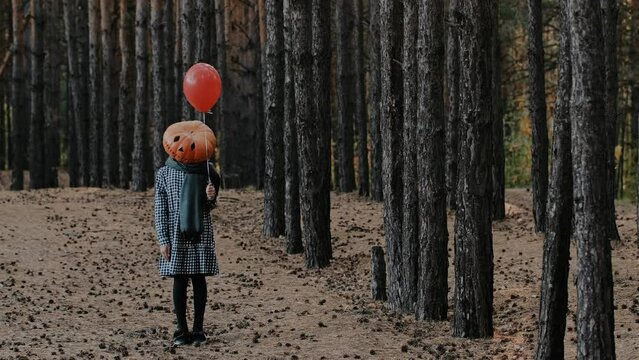 A teenage girl in a giant pumpkin head stands in the woods with a red ball in her hands and looks into the camera. Halloween concept