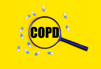 Pharmaceutical tablets and capsules near a magnifying glass with the text COPD Chronic Obstructive Pulmonary Disease on a yellow background. View from above.