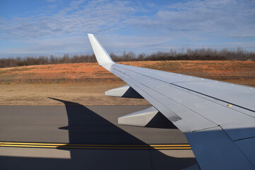 View from an airplane window of a wing and its shadow. Plane on runway ready to take off, with blue sky and cloudy background.