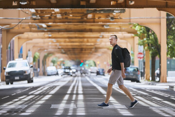 Side view of adult man with backpack walking on pedestrian crosswalk uder elevated railway of public transportation. Chicago, United States. - Powered by Adobe