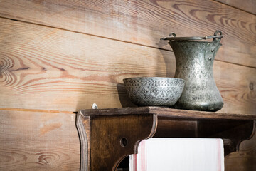 Traditional old metal vessels from the early 19th century standing on a shelf against a wooden wall. A linen towel hangs from a wooden shelf. Background in retro style.