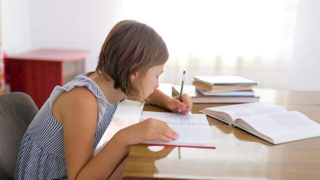 Side view school child girl student carefully, concentrate writes in notebook while sitting at table. Child is left hander, short hair, there are textbooks on table. Hard work doesn't let relax.
