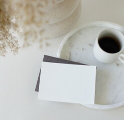 Blank paper sheet card mockup in  breakfast still life scene.Cup of coffee on tray, grass in a vase top view on white table. Copy space.Feminine lifestyle composition. 
