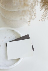 Blank paper sheet card mockup on marble tray and grass in a vase top view on white table. Copy space.Feminine lifestyle composition. 