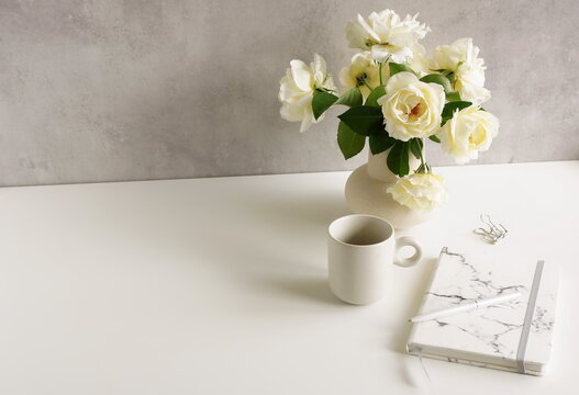 Feminine lifestyle composition, minimal workspace neutral colors. Vase with beige roses flowers , cup coffee,diary,pen on white desk near  gray background with copy space .Breakfast stationary mockup.