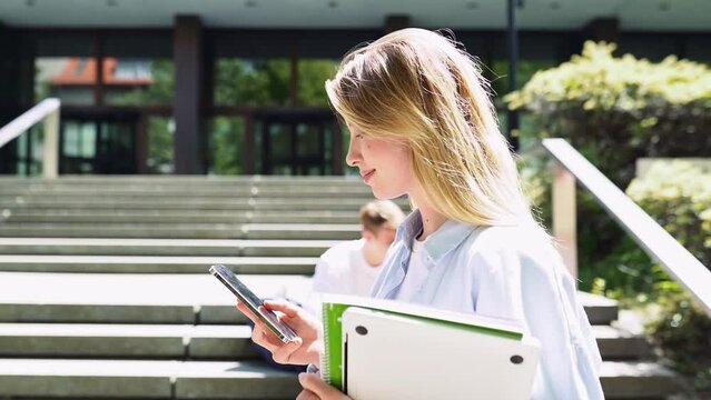 Pretty blonde girl student social media user holding cell phone using mobile applications tech on smartphone looking at cellphone, chatting, texting, walking outside campus in slow motion, side view.