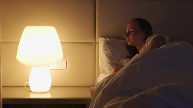 Woman wakes up at midnight switching on bright lamp on bedside table to illuminate hotel room. Female tourist turns on light lying in bed closeup