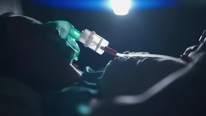 A female patient in medical mask is breathing oxygen support in a hospital next to heart rate machine. Unhealthy woman in coma. Getting mechanical ventilation. Concept of pandemic and therapy.