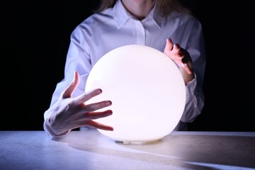 Businesswoman using glowing crystal ball to predict future at table in darkness, closeup. Fortune...