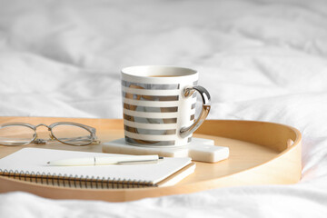 Mug of hot drink with stylish cup coaster, glasses and notebook on bed in room