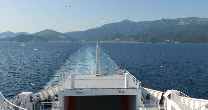 Summer seascape with seagulls flying around ferry. Sea water waves trails from ferry boat. View from the ferries. Sunny clear blue sky. Mountain landscape of Thassos, Greece.