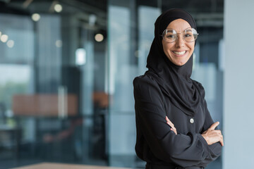 Portrait of muslim woman in hijab at work in office, business woman smiling and looking at camera with crossed arms. - obrazy, fototapety, plakaty