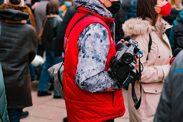 Fototapeta na wymiar A correspondent in a red jacket with a camera working in the crowd. Demonstration. Film. Journalism. Politic. Against. Street. Record. Broadcasting. Revolution. Cam. Reporter. Reportage. March