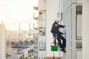 Industrial mountaineering worker in uniform hangs over residential building at city background,...