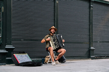 young man musician with beard looking at camera and playing accordion in a tourist street in...