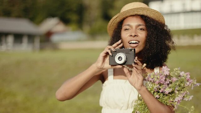 4k Portrait of attractive african-american woman in hat making photos on retro camera in the summer field. Countryside and farmland style.