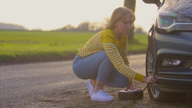 Woman inflating flat car tyre with electric pump on country road - shot in slow motion