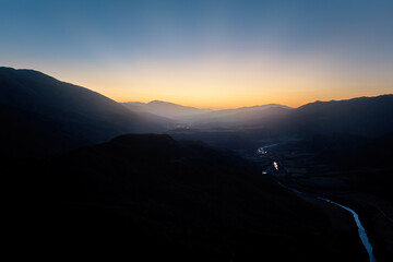 Aerial panoramic view of the dark valley of the Vjose River after sunset, surrounded by the silhouettes of majestic mountains. Orange-blue sky, sun rays behind the mountains, summer. 