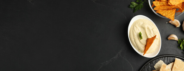Delicious hummus and tortilla chips on black table, flat lay with space for text. Banner design