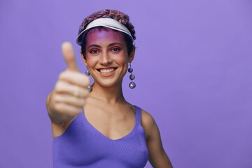 Portrait of a sporty fashion woman posing smiling with teeth and pointing a finger at the camera in a purple yoga tracksuit and a transparent cap on a purple monochrome background