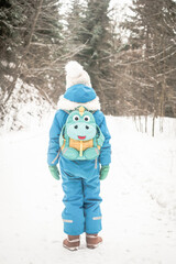 Six years old child, boy standing with his back to camera. Kid with funny backpack in winter forest full of snow. Colorfully dressed child. Backpacking in winter condition in the forest. 