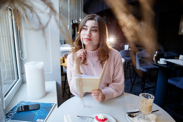 Satisfied woman in sweater enjoying day in modern cafe with cup of hot drink and writing in diary.