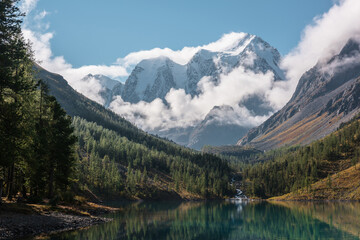 Naklejka na ściany i meble Scenic landscape with turquoise mountain lake in autumn valley against large snow mountains in low clouds in morning sunlight. Alpine lake with view to sunlit high snowy mountain range in low clouds.