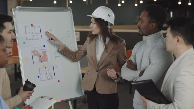 Multicultural team of experienced architects discussing project of building at modern office. Beautiful woman in helmet standing near flip chart and explaining peculiarities of new design.