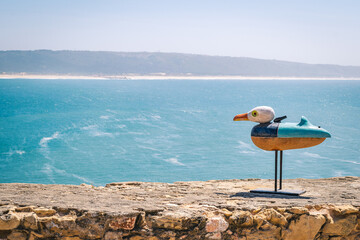 A bird statue on a stand on a cliff overlooking the ocean and a beach with a mountain in the background