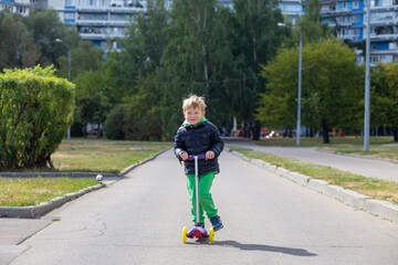 A blond boy 3-4 years old in a black jacket and green trousers rides a scooter along an alley in the city.