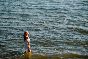Woman in summer dress standing on seashore and looking at horizon. Young beautiful girl standing in water