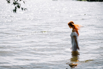 Woman in summer white dress stands on the seashore and looks at the horizon. Young beautiful girl standing in the water