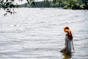 Woman in summer white dress stands on the seashore and looks at the horizon. Young beautiful girl standing in the water