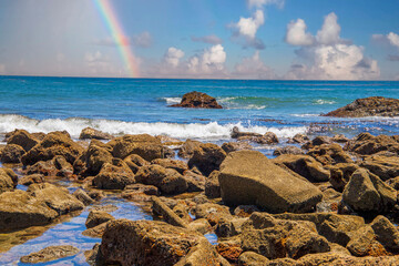 a gorgeous summer landscape at the beach with vast deep blue ocean water and large rocks along the shore with waves rolling over the rocks with blue sky, clouds and a rainbow at Royal Beach park