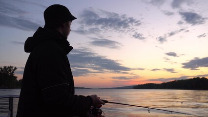 A fisherman in a jacket is fishing at sunset on the river from a boat. A young man throws a bait on...