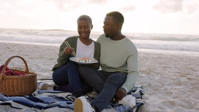 Sushi, beach and picnic couple dating or on romantic honeymoon date with ocean, sea and sky. Happy, love and romance woman and man or black people eating food together for valentines day anniversary