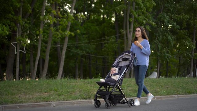 Mom walks with a baby in a stroller and eats a burger on the go. A young woman in glasses pushes a stroller with her son and eats a burger in the park.